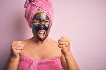 Middle age brunette woman wearing beauty black face mask over isolated pink background angry and mad raising fists frustrated and furious while shouting with anger. Rage and aggressive concept.