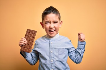 Young little boy kid eating sweet chocolate bar for dessert over isolated yellow background annoyed...
