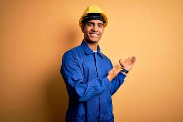 Young handsome african american worker man wearing blue uniform and security helmet Inviting to enter smiling natural with open hand