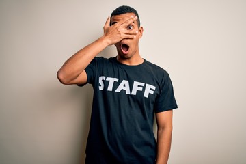 Young handsome african american worker man wearing staff uniform over white background peeking in shock covering face and eyes with hand, looking through fingers with embarrassed expression.