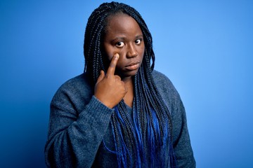 African american plus size woman with braids wearing casual sweater over blue background Pointing to the eye watching you gesture, suspicious expression