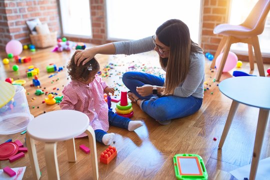 Young beautiful teacher and toddler sitting on the floor building pyramid using hoops at kindergarten