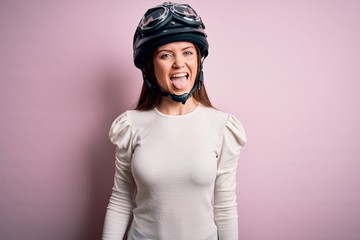 Young beautiful motorcyclist woman with blue eyes wearing moto helmet over pink background sticking tongue out happy with funny expression. Emotion concept.