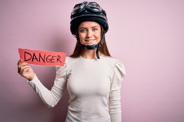 Beautiful motorcyclist woman with blue eyes wearing moto helmet holding danger message with a happy face standing and smiling with a confident smile showing teeth