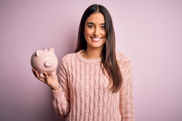 Young beautiful brunette woman holding piggy bank saving money for retirement with a happy face...