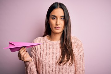 Young beautiful brunette woman holding paper airplane over isolated pink background with a...