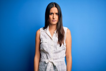 Young beautiful brunette woman wearing casual dress over isolated blue background skeptic and...