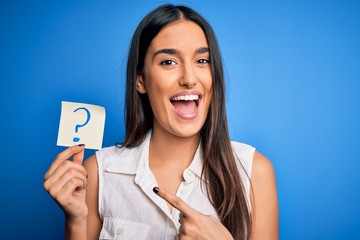 Young beautiful brunette woman holding paper with question mark symbol message very happy pointing with hand and finger