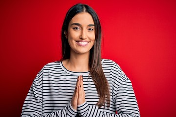 Young beautiful brunette woman wearing casual striped t-shirt over red background praying with hands together asking for forgiveness smiling confident.