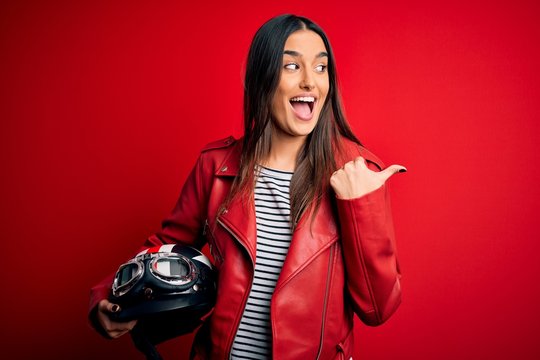 Young beautiful brunette motorcyclist woman holding motorcycle helmet and red jacket pointing and showing with thumb up to the side with happy face smiling