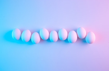 White chicken eggs aligned in a row on empty plain background lit with blue and pink gel filter lights. Creative minimalist style trendy Easter concept.
