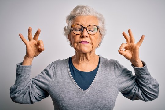 Senior beautiful grey-haired woman wearing casual sweater and glasses over white background relax and smiling with eyes closed doing meditation gesture with fingers. Yoga concept.