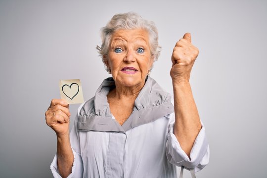 Senior beautiful grey-haired woman holding reminder paper heart over white background annoyed and frustrated shouting with anger, crazy and yelling with raised hand, anger concept