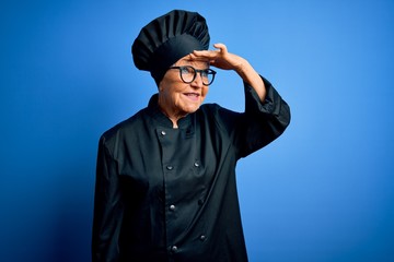 Senior beautiful grey-haired chef woman wearing cooker uniform and hat over blue background very happy and smiling looking far away with hand over head. Searching concept.