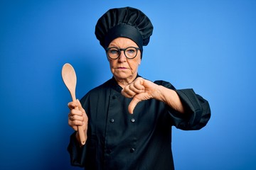 Naklejka premium Senior beautiful grey-haired chef woman wearing cooker uniform and hat holding wooden spoon with angry face, negative sign showing dislike with thumbs down, rejection concept