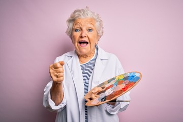 Senior beautiful grey-haired artist woman painting using brush and palette over pink background screaming proud and celebrating victory and success very excited, cheering emotion - obrazy, fototapety, plakaty