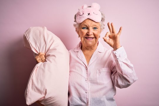 Senior beautiful woman wearing sleep mask holding pillow over isolated pink background doing ok sign with fingers, excellent symbol