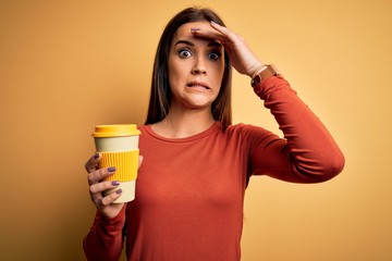 Young beautiful brunette woman drinking cup of takeaway coffe over yellow background stressed with hand on head, shocked with shame and surprise face, angry and frustrated. Fear and upset for mistake.