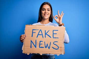 Young beautiful brunette woman holding banner with fake news message doing ok sign with fingers,...