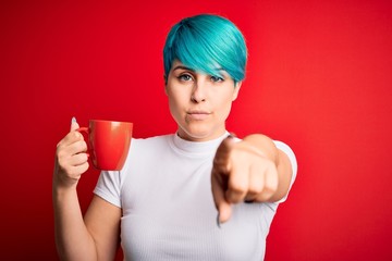 Young woman with blue fashion hair drinking a cup of coffee s over red isolated background pointing with finger to the camera and to you, hand sign, positive and confident gesture from the front