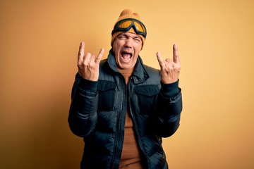 Middle age handsome grey-haired skier man on vacation wearing ski goggles shouting with crazy expression doing rock symbol with hands up. Music star. Heavy concept.