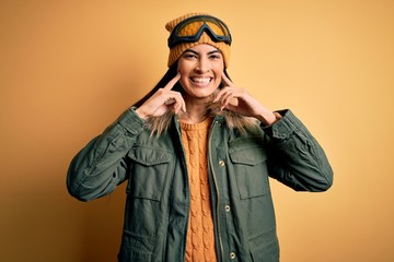 Young beautiful hispanic woman wearing ski glasses and coat for winter weather Smiling with open mouth, fingers pointing and forcing cheerful smile
