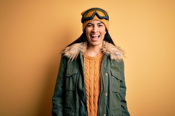 Young beautiful hispanic woman wearing ski glasses and coat for winter weather winking looking at the camera with sexy expression, cheerful and happy face.