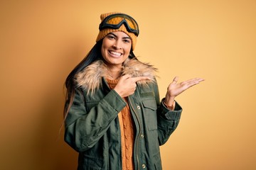 Young beautiful hispanic woman wearing ski glasses and coat for winter weather amazed and smiling to the camera while presenting with hand and pointing with finger.