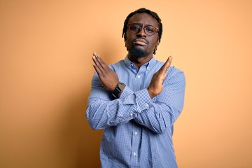 Young handsome african american man wearing shirt and glasses over yellow background Rejection...