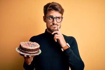 Young man with beard wearing glasses holding birthday cake over isolated yellow background serious...