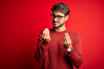 Young handsome man with beard wearing glasses and sweater standing over red background doing money...