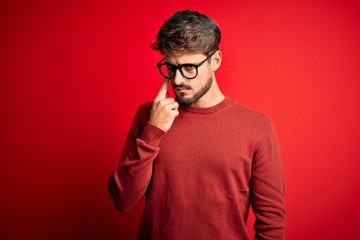 Young handsome man with beard wearing glasses and sweater standing over red background Pointing to the eye watching you gesture, suspicious expression