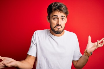 Young handsome man with beard wearing casual t-shirt standing over red background clueless and confused expression with arms and hands raised. Doubt concept.