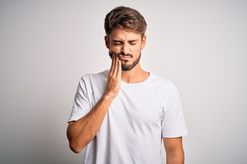 Young handsome man with beard wearing casual t-shirt standing over white background touching mouth with hand with painful expression because of toothache or dental illness on teeth. Dentist