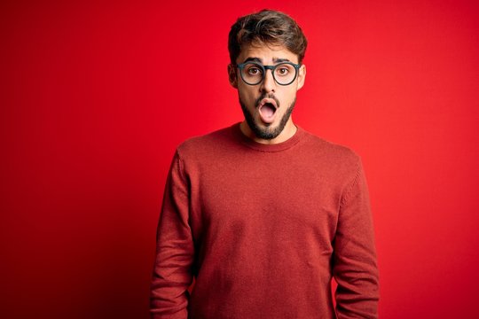 Young handsome man with beard wearing glasses and sweater standing over red background afraid and shocked with surprise and amazed expression, fear and excited face.