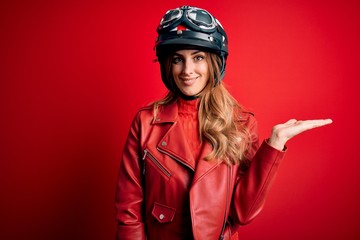 Young beautiful brunette motrocyclist woman wearing moto helmet over red background smiling cheerful presenting and pointing with palm of hand looking at the camera.