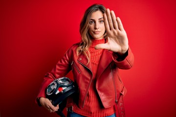 Beautiful brunette motorcyclist woman wearing red jacket holding motorcycle helmet with open hand doing stop sign with serious and confident expression, defense gesture