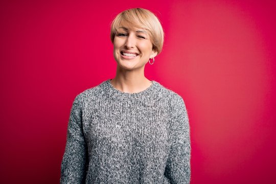 Young blonde woman with modern short hair wearing casual sweater over pink background winking looking at the camera with sexy expression, cheerful and happy face.