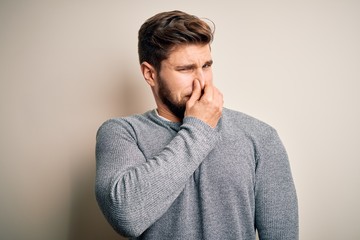 Young handsome blond man with beard and blue eyes wearing casual sweater smelling something stinky and disgusting, intolerable smell, holding breath with fingers on nose. Bad smell