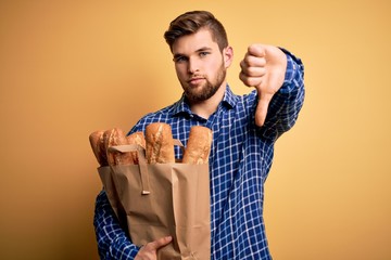 Young blond man with beard and blue eyes holding paper bag with bread with angry face, negative...
