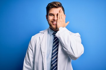 Young blond therapist man with beard and blue eyes wearing coat and tie over background covering one eye with hand, confident smile on face and surprise emotion.
