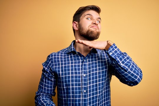 Young blond businessman with beard and blue eyes wearing shirt over yellow background cutting throat with hand as knife, threaten aggression with furious violence