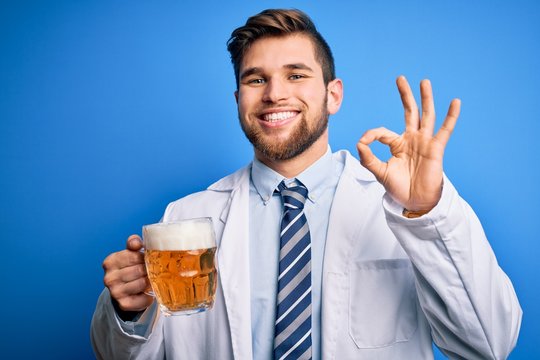 Young blond doctor man with beard and blue eyes wearing coat drinking jar of beer doing ok sign with fingers, excellent symbol