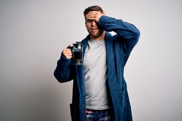 Fototapeta na wymiar Young blond man with beard and blue eyes wearing pajama making coffe using coffeemaker stressed with hand on head, shocked with shame and surprise face, angry and frustrated. Fear and upset