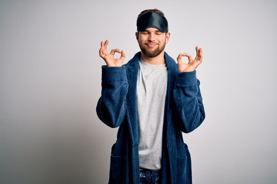 Young blond man with beard and blue eyes wearing sleep mask and pajama relax and smiling with eyes closed doing meditation gesture with fingers. Yoga concept.