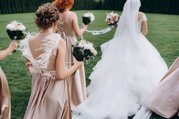 Rear view of elegant bridesmaids walk on the green lawn on outdoors and holding beautiful modern a...