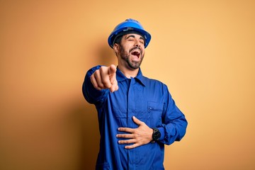 Mechanic man with beard wearing blue uniform and safety helmet over yellow background laughing at...