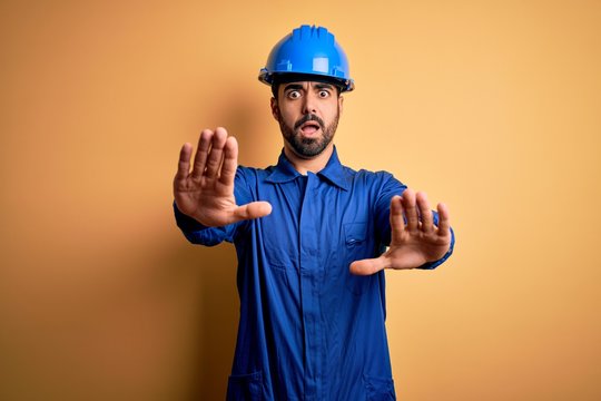 Mechanic man with beard wearing blue uniform and safety helmet over yellow background doing stop gesture with hands palms, angry and frustration expression