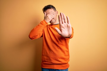 Young handsome man wearing orange casual sweater standing over isolated yellow background covering eyes with hands and doing stop gesture with sad and fear expression. Embarrassed and negative