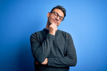 Young handsome man wearing casual sweater and glasses standing over blue background looking confident at the camera with smile with crossed arms and hand raised on chin. Thinking positive.
